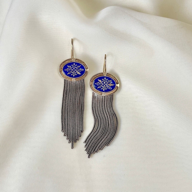 K&ouml;nnte beinhalten: Ein Paar Ohrh&auml;nger mit ovalem, blauem Emaille-Design und silberfarbener Kettenfransen. Die Ohrringe haben einen goldfarbenen Rahmen und Haken. Das Design auf dem blauen Emaille ist ein wei&szlig;es Blumenmuster.