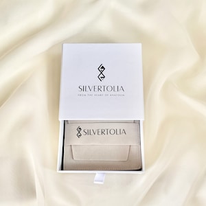 Puede incluir: Una caja de joyer&iacute;a blanca con un interior beige. La caja tiene el logotipo "SILVERTOLIA from the heart of Anatolia" impreso en la parte superior. El interior de la caja tiene el logotipo "SILVERTOLIA" impreso.