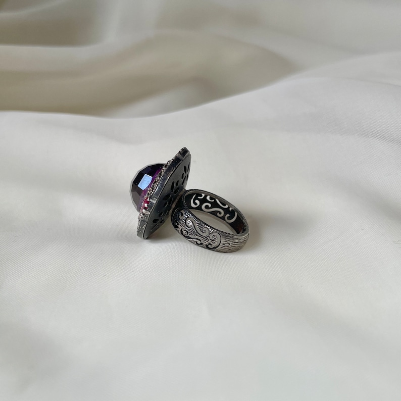 Puede incluir: Un anillo de plata con una gran piedra preciosa p&uacute;rpura facetada engastada en un dise&ntilde;o calado detallado. La banda del anillo tambi&eacute;n est&aacute; dise&ntilde;ada de forma intrincada con un acabado texturizado.