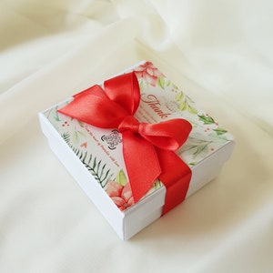 K&ouml;nnte beinhalten: Eine wei&szlig;e Geschenkbox mit floralem Design und einem leuchtend roten Band, das zu einer Schleife gebunden ist. Auf der Box steht "Thank" und "SILVERFOLIA". Die Box befindet sich auf einem wei&szlig;en Stoffhintergrund.