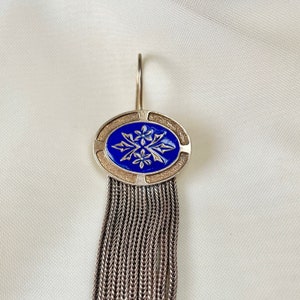K&ouml;nnte beinhalten: Silberner Ohrring mit blauem, ovalem Mittelst&uuml;ck mit floralem Design. Das Oval ist von einem strukturierten Rand umgeben und h&auml;ngt an einem Haken. Unterhalb des Ovals sorgt eine Kaskade aus silbernen Ketten f&uuml;r Bewegung.