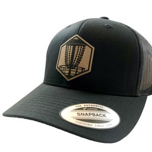 Puede incluir: Gorra de camionero negra con un parche hexagonal marrón que presenta un diseño de cesta de disc golf. La gorra tiene una visera curva y una parte trasera de malla. El parche dice "THE AUTHENTIC SNAPBACK YUPOONG SINCE 1974".