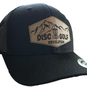 May include: Black trucker hat with a mesh back and a curved brim. The front features a tan leather patch with a mountain and disc golf basket graphic, and the words "DISC GOLF REVOLUTION".