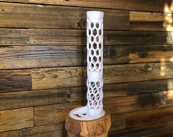 May include: A white, 3D-printed decorative object with a honeycomb pattern. The object is cylindrical with a circular base and is displayed on a wooden surface. The design features open hexagonal shapes throughout its structure.