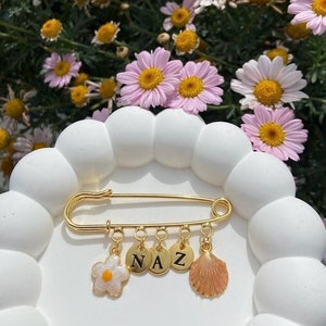 May include: Gold safety pin charm with a white flower, three gold circles with the letters N, A, and Z, and a gold seashell charm. The charm is on a white background.