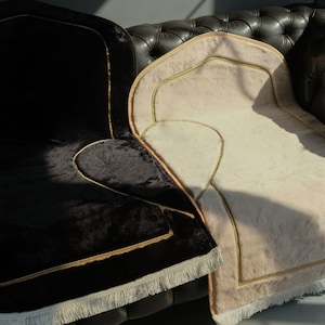 Puede incluir: Dos alfombras de oración con ribetes dorados y flecos. Una alfombra es de terciopelo negro y la otra es beige.