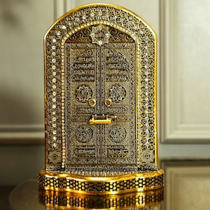 Könnte beinhalten: Eine goldfarbene Metallreplik der Kaaba, das kubische Gebäude im Zentrum der heiligsten Moschee des Islam, der Masjid al-Haram in Mekka, Saudi-Arabien. Die Replik ist mit komplizierten Details verziert und verfügt über eine schwarze Tür mit arabischer Kalligraphie.