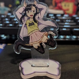 May include: Anime-style acrylic standee featuring a girl with glasses, wearing a yellow shirt with a black cat graphic and the text "SUGAR RUSH". She's in a white jacket and pink skirt. The stand has a bone-shaped base.