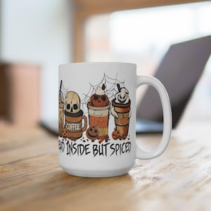 Może przedstawiać: Biały ceramiczny kubek do kawy z wzorem o tematyce Halloween przedstawiającym cztery różne napoje kawowe ze straszną twarzą. Na kubku nadrukowany jest tekst "Dead inside but spiced".