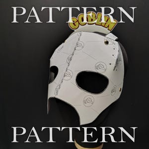 May include: A white paper mask template featuring a repeating circular icon pattern and the word GOBLIN printed at the top. The mask has two large eye openings and is constructed with small metal fasteners along the seams and edges.