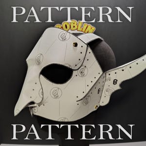 May include: A white mask with a pointed nose and a wing-like side extension, featuring a repeating black line art pattern of thumbs-up icons and the letter B. The mask is constructed from multiple panels joined by small gold-coloured metal rivets.
