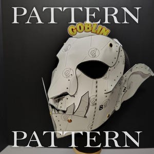 May include: A white paper goblin mask featuring a repeating thumbs-up icon pattern, prominent pointed ears, and large tusks. The mask is assembled with small gold-colored metal fasteners and displays the word GOBLIN in yellow and orange text.