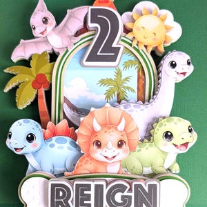 Op de afbeelding: Een kleurrijke taarttopper met dinosaurus thema met het nummer 2, een vulkaan en een lachende zon. De topper heeft verschillende cartoon dinosaurussen, palmbomen en de naam "REIGN" op een botvormige banner.