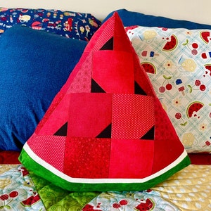 May include: A red and green watermelon-shaped cushion with a white rind. The cushion is made of patchwork fabric and is sitting on a bed with a floral patterned quilt.