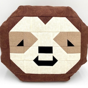 May include: A brown and tan patchwork pillow with a sloth face design. The sloth's eyes are black triangles, and its nose and mouth are black.