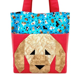 Pode incluir: Uma sacola vermelha com bolinhas brancas e alças vermelhas. A sacola apresenta um rosto de cachorro em patchwork na frente e um forro azul com vários padrões de raças de cães e ossos.