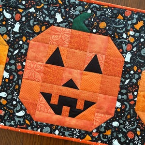 May include: A rectangular quilted table runner featuring a central orange jack-o'-lantern block made of various orange fabric squares. The border is a black fabric printed with white ghosts, orange pumpkins, black cats, and keys, finished with an orange binding.