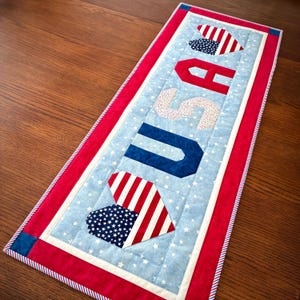 May include: A rectangular quilted table runner featuring a light blue fabric with white stars. The center displays the letters USA in red, white, and blue patterns, flanked by two heart shapes. The border is red with a blue and white striped edge.