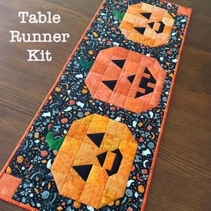 May include: A rectangular quilted table runner kit featuring three orange pumpkin blocks with black geometric facial features. The background fabric is black with a pattern of small white ghosts, orange pumpkins, and green leaves. The text Table Runner Kit is visible.