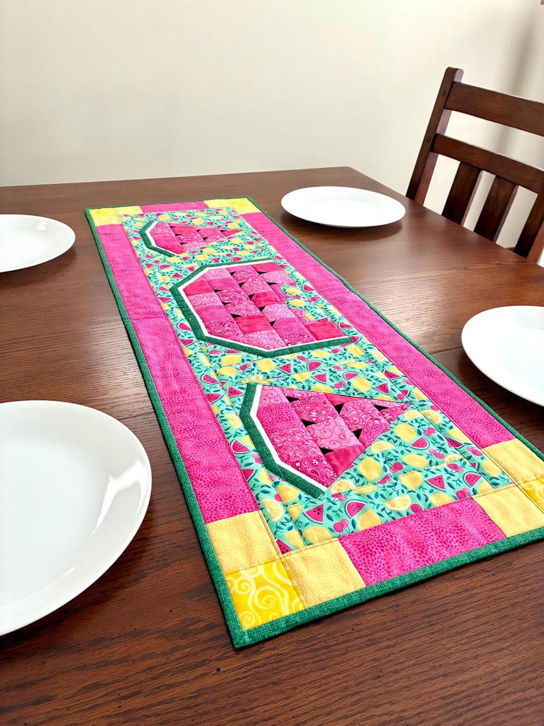 May include: A rectangular quilted table runner featuring three watermelon slice motifs. The design includes pink, yellow, and green fabric patches with a repeating pattern of small watermelons and lemons on a light blue background, finished with a green border.