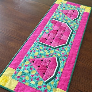 May include: A quilted table runner with a watermelon theme. The runner features pink watermelon slices, a turquoise background with lemons and watermelon slices, and a pink border. The runner is approximately 30 cm wide and 91 cm long.