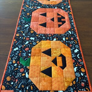 May include: A rectangular quilted table runner featuring three pieced orange pumpkin blocks with black triangular eyes and mouths. The background fabric is black with a repeating pattern of ghosts, owls, keys, and pumpkins, finished with an orange border.