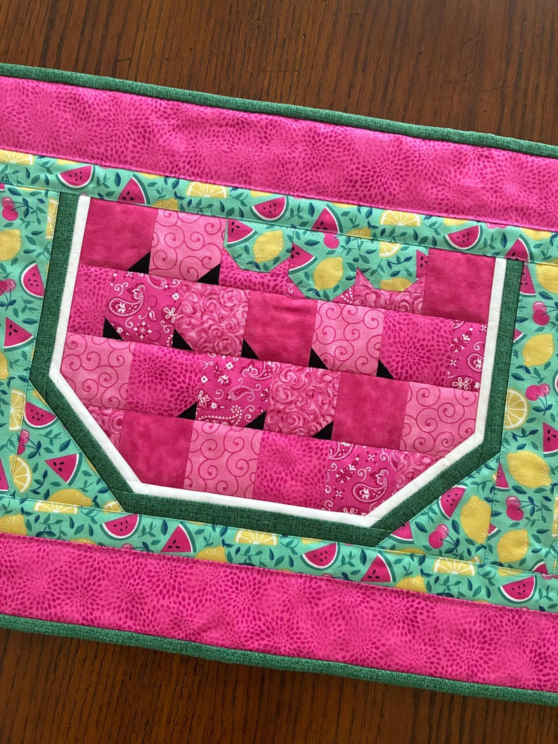 K&ouml;nnte beinhalten: Gesteppter Tischl&auml;ufer mit Wassermelonen-Design. Die Mitte zeigt eine rosa Wassermelonenscheibe mit wei&szlig;er Schale und schwarzen Kernen. Der L&auml;ufer hat einen rosa Rand und einen gr&uuml;n-t&uuml;rkisfarbenen Hintergrund mit Wassermelonen- und Zitronenmustern.