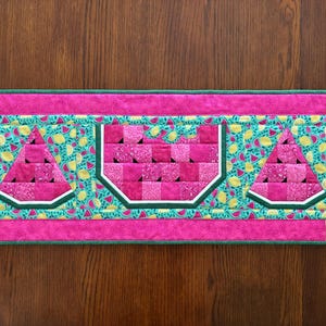 K&ouml;nnte beinhalten: Ein rechteckiger, gesteppter Tischl&auml;ufer mit Wassermelonen-Design. Der L&auml;ufer hat einen rosa Rand und eine t&uuml;rkisfarbene Mitte mit Wassermelonen- und Zitronenmustern. Drei Wassermelonenscheiben sind deutlich zu sehen.