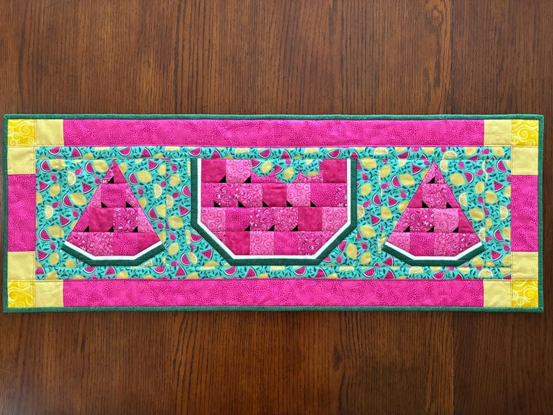 K&ouml;nnte beinhalten: Ein rechteckiger, gesteppter Tischl&auml;ufer mit Wassermelonen-Design. Der L&auml;ufer hat einen rosa Rand und eine t&uuml;rkisfarbene Mitte mit Wassermelonen- und Zitronenmustern. Drei Wassermelonenscheiben sind deutlich zu sehen.