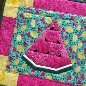 K&ouml;nnte beinhalten: Ein gesteppter Tischset mit einem Wassermelonen-Scheiben-Design. Der Tischset hat einen gr&uuml;nen Rand mit rosa und gelben Stoffakzenten mit Muster. Die Wassermelonenscheibe besteht aus rosa Stoff mit schwarzen Kerndetails, vor einem t&uuml;rkisfarbenen Hintergrund mit Fr&uuml;chten.