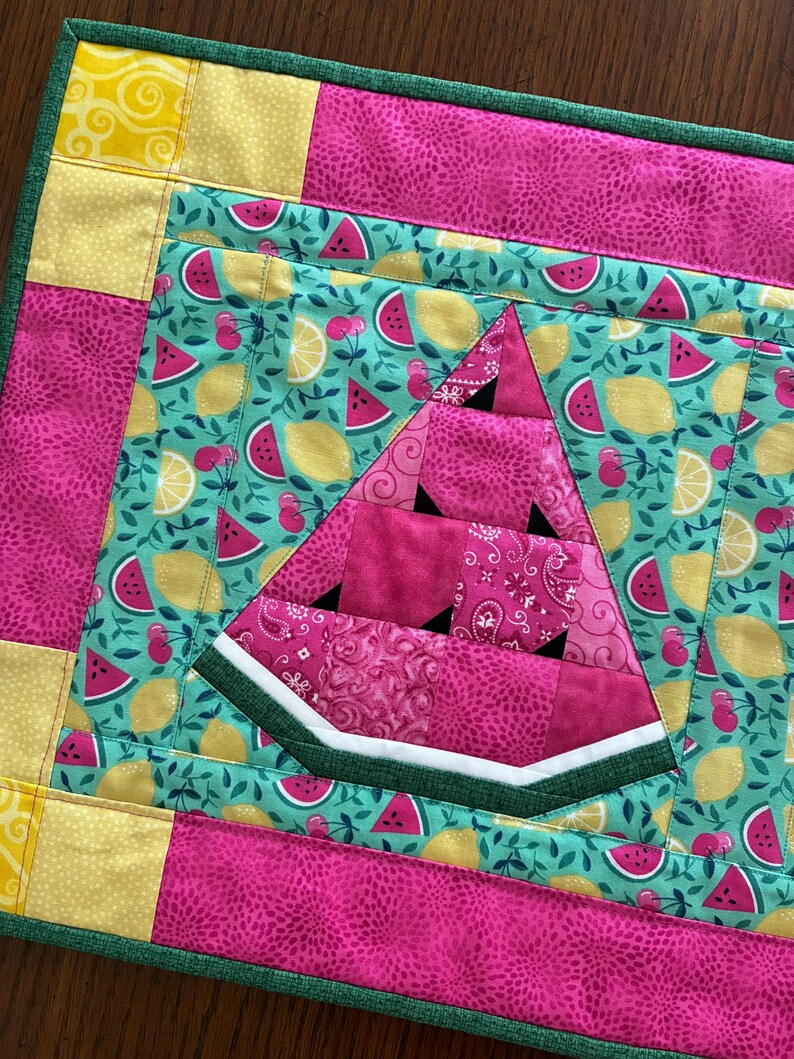 K&ouml;nnte beinhalten: Ein gesteppter Tischset mit einem Wassermelonen-Scheiben-Design. Der Tischset hat einen gr&uuml;nen Rand mit rosa und gelben Stoffakzenten mit Muster. Die Wassermelonenscheibe besteht aus rosa Stoff mit schwarzen Kerndetails, vor einem t&uuml;rkisfarbenen Hintergrund mit Fr&uuml;chten.