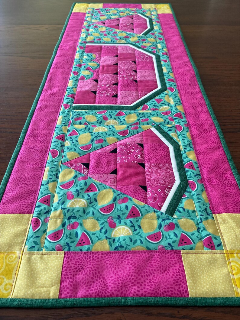 K&ouml;nnte beinhalten: Ein rechteckiger, gesteppter Tischl&auml;ufer mit einem fruchtigen Muster. Der L&auml;ufer zeigt eine Kombination aus rosa, gelben und t&uuml;rkisfarbenen Stoffen. Das Design umfasst Wassermelonen-, Zitronen- und Kirschmotive mit geometrischen Formen und Bord&uuml;ren.