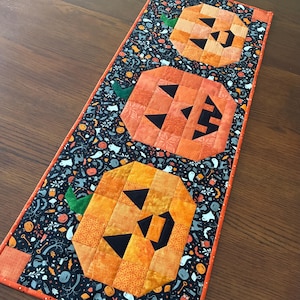 Könnte beinhalten: Ein Halloween-Tischläufer mit drei orangefarbenen, gesteppten Kürbisgesichtern mit schwarzen Dreiecksaugen und -mündern. Die Kürbisse befinden sich auf einem schwarzen Hintergrund mit einem sich wiederholenden Halloween-Muster. Der Läufer ist mit orangefarbenem Stoff eingefasst.