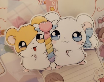May include: A sticker featuring two cartoon hamsters with large eyes. One hamster is tan and the other is white. They are holding a rainbow wand with a heart on top. The background includes stationery with pink and purple accents.