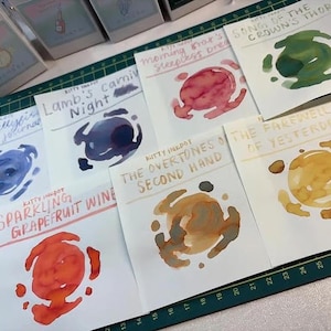 May include: A collection of white paper swatches featuring ink samples in various colors including blue, dark purple, pink, green, orange, brown, and yellow. Each swatch displays the brand name Kitty Inkpot and a unique handwritten title.