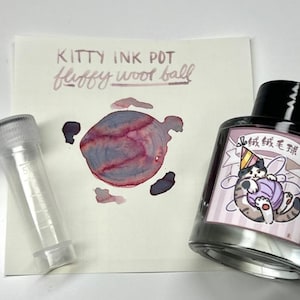 May include: A glass ink bottle with a black cap and a label featuring a cat wearing a party hat and holding a ball of yarn. Next to it is a clear 5ml plastic vial and a paper swatch showing the ink color as a blend of purple and pink.