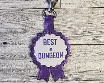 Op de afbeelding: Een paarse sleutelhanger in de vorm van een lint met een wit cirkelvormig midden waarop de tekst BEST in DUNGEON in het paars is gedrukt. Het item bevat een zilverkleurige metalen karabijnhaak aan de bovenste lus voor bevestiging.
