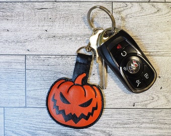 May include: A black leather pumpkin keychain with a scary face. The keychain is attached to a silver key ring with a car key.