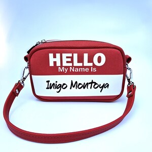 May include: A red crossbody bag with a white name tag design. The bag features the text "HELLO My Name Is" above the name "Inigo Montoya" in a handwritten font. The bag has a red strap and silver hardware.
