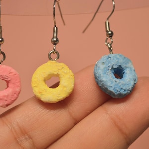 Puede incluir: Primer plano de tres pendientes con colgantes circulares de colores. Los colgantes son rosas, amarillos y azules, que se asemejan a cereales. Cada colgante está suspendido de un gancho plateado.