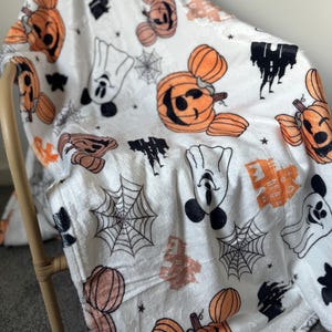 Op de afbeelding: Witte fleece deken met een Halloween-thema. De deken heeft Mickey Mouse en pompoenen met een zwart-wit spinnenwebpatroon.