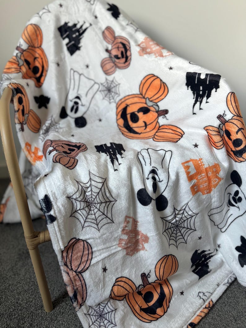 Op de afbeelding: Witte fleece deken met een Halloween-thema. De deken heeft Mickey Mouse en pompoenen met een zwart-wit spinnenwebpatroon.