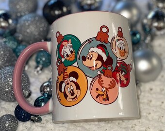 Op de afbeelding: Witte keramische mok met een roze handvat met een kerstontwerp van Mickey Mouse, Minnie Mouse, Donald Duck, Goofy en Pluto als kerstversiering.