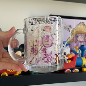 May include: A clear glass mug featuring a design of various magical items, including a potion bottle, a skull, a wand, a moth, a candle, and a spine. The text "Defense Against The Dark Arts" is printed on the mug.