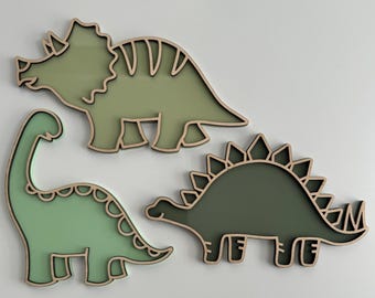 Könnte beinhalten: Drei hölzerne Dinosaurier-Wanddekorationen in Grün und Braun. Die Dinosaurier sind ein Triceratops, ein Stegosaurus und ein Brachiosaurus.