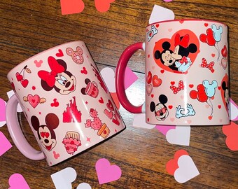 Op de afbeelding: Twee roze en rode keramische mokken met een Valentijnsdagthema. De ene mok heeft Mickey en Minnie Mouse, de andere heeft Mickey en Minnie Mouse met harten, kersen en andere Valentijnsdagsymbolen.