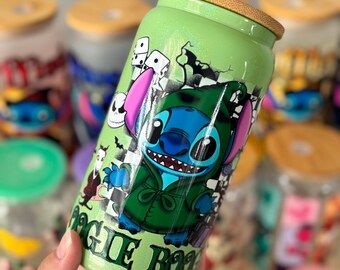 Op de afbeelding: Een groene glazen beker met een bamboe deksel met een cartoon karakter ontwerp. Het ontwerp bevat het karakter in een groene outfit met de tekst "MAGIC BOO". Andere bekers zijn zichtbaar op de achtergrond.
