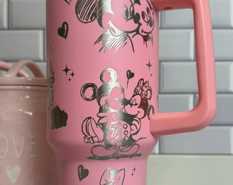 Op de afbeelding: Roze geïsoleerde beker met een zilveren Mickey en Minnie Mouse ontwerp. De beker heeft een handvat, een doorzichtig deksel en een roze klapdeksel. Het ontwerp bevat harten en sterren.