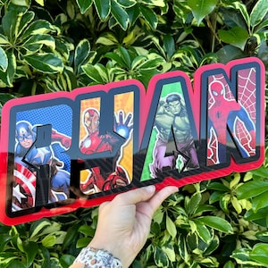 May include: A wooden sign spelling "RYAN" with each letter showcasing a Marvel superhero: Captain America, Iron Man, Hulk, and Spider-Man. The sign has a red border and is set against a background of green leaves.