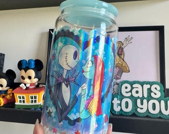 Op de afbeelding: Een doorzichtige glazen beker met een lichtblauw deksel en een holografische sticker van Jack Skellington en Sally uit The Nightmare Before Christmas. De personages staan in een groot blauw hart dat omringd is door kleine sterretjes.